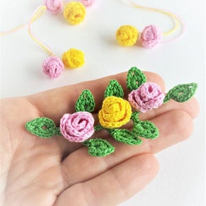 Puede incluir: Primer plano de rosas de ganchillo hechas a mano en rosa y amarillo con hojas verdes. Varias rosas y capullos individuales están dispersos sobre una superficie blanca. Las rosas se sostienen en una mano, mostrando la delicada artesanía y los colores vibrantes.