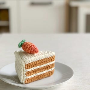 May include: A crocheted slice of carrot cake with white icing and a small orange carrot on top. The cake is on a white plate, highlighting the handmade details and the playful design. The cake is a charming decorative item.