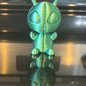 May include: A 3D-printed, teal-colored alien figurine with large oval eyes, small arms, and antennae. The alien is sitting, with its reflection visible below. The figurine is made of a shiny, textured material.