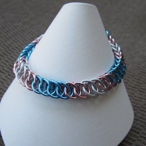 May include: A blue, white, and pink chainmail bracelet. The bracelet is made of small metal rings that are linked together.