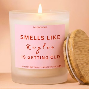 May include: A lit, frosted glass candle with a pink label that reads "Smells Like Kaylee Is Getting Old." The candle has a wooden lid and is labeled as 8 oz soy wax. The background is a soft peach color.