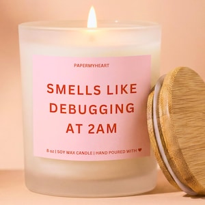 May include: A lit, frosted glass candle with a wooden lid. The candle has a pink label with the text "SMELLS LIKE DEBUGGING AT 2AM". The candle is 8 oz and made of soy wax.