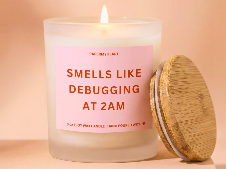 May include: A lit, frosted glass candle with a wooden lid. The candle has a pink label with the text "SMELLS LIKE DEBUGGING AT 2AM". The candle is 8 oz and made of soy wax.