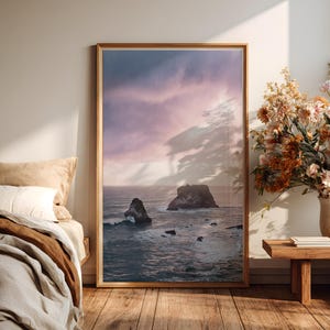Könnte beinhalten: Ein gerahmter Kunstdruck zeigt eine Küstenlandschaft mit Meereswellen und Felsformationen unter einem dramatischen Himmel. Das Kunstwerk wird neben einem Bett mit neutraler Bettwäsche und einem hölzernen Beistelltisch mit einer Blumenvase präsentiert.