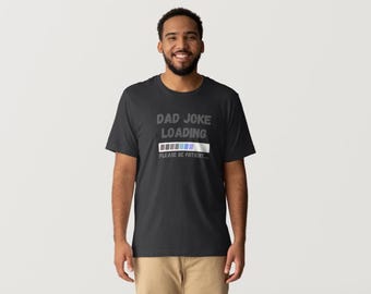 Puede incluir: Una camiseta negra con el texto "DAD JOKE LOADING" sobre una barra de carga. El texto "PLEASE BE PATIENT" está debajo. La camiseta la lleva una persona.