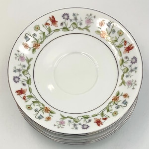 May include: A stack of white ceramic saucers with a floral design. The saucers have a gold rim and a border of colorful flowers and green vines. The flowers are red, orange, purple, and pink.