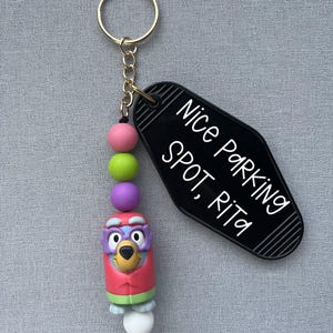 Puede incluir: Un llavero con un anillo y cadena dorados, con una etiqueta negra estilo motel con el texto "Nice Parking Spot, Rita". El llavero también incluye cuentas de colores y un dije de personaje de dibujos animados.