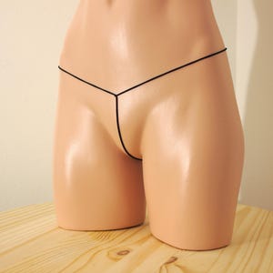 May include: Black thong underwear displayed on a mannequin. The underwear is made of a thin, black material and features a minimalist design. The mannequin is a light skin tone and is positioned on a wooden surface.
