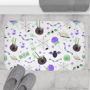 May include: A rectangular white bath mat featuring a Halloween pattern. The design includes purple, green, and black illustrations of cauldrons, spiders, cobwebs, candy, and other spooky elements. Grey slippers are in the foreground.
