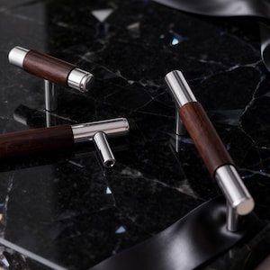 Könnte beinhalten: Drei Schrankgriffe mit einem dunklen Holzmittelteil und silberfarbenen Metallenden und -pfosten werden auf einer schwarzen Marmoroberfläche präsentiert. Die Griffe sind so angeordnet, dass sie ihr Design und ihre Handwerkskunst zeigen.