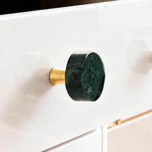 Może przedstawiać: Zbliżenie białej szuflady z dwoma zielonymi marmurowymi i złotymi gałkami do szafek. Gałki są cylindryczne z ciemnozieloną, żyłkowaną marmurową powierzchnią i złotą podstawą. Szuflada jest otwarta.
