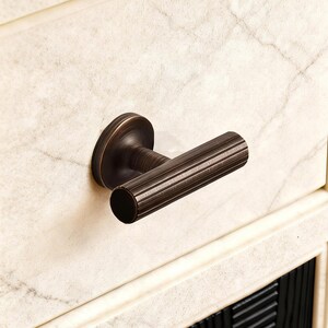 May include: Bronze cabinet handle with a cylindrical, ridged grip and a circular backplate. The handle is mounted on a cream-colored cabinet with a marble-like pattern. The overall design suggests a vintage or antique aesthetic.