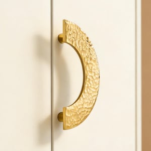 May include: A gold-colored cabinet pull with a textured, hammered finish. The curved handle is mounted on a white cabinet door. The hardware has a semi-circular design and a modern aesthetic.