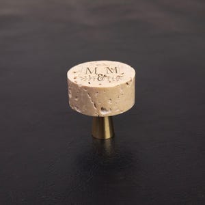 Könnte beinhalten: Ein runder, hellbrauner Korkknopf mit einer flachen Oberseite, die mit dem eingravierten Text M & M und kleinen Blattmotiven versehen ist. Der Korkaufsatz sitzt auf einem glatten, konischen, goldfarbenen Metallsockel, der nach unten zuläuft.