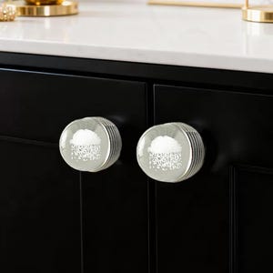 May include: Two clear glass cabinet knobs with a white cloud design and silver ribbed edges. The knobs are mounted on a black cabinet door. The background shows a white countertop. Decorative hardware.
