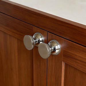 May include: Close-up of two polished chrome cabinet knobs with a round, flat design. The knobs are mounted on a dark brown wooden cabinet with visible wood grain. The cabinet is part of a larger piece of furniture.