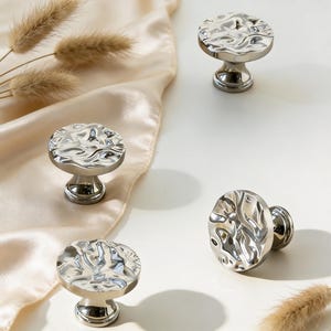 May include: Four silver cabinet knobs with a textured, rippled surface are arranged on a white background with a beige satin fabric. The knobs have a round top and a pedestal base. Dried ornamental grass is also present.