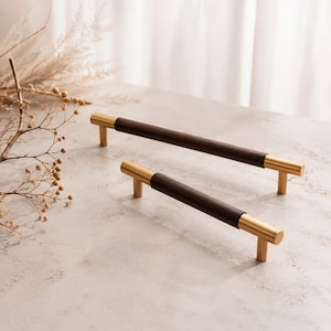 May include: Two cabinet pulls featuring a cylindrical dark brown wood centre section flanked by brushed gold metal ends and mounting posts. The hardware pieces are arranged diagonally, showcasing a smooth finish and a straight, linear design.
