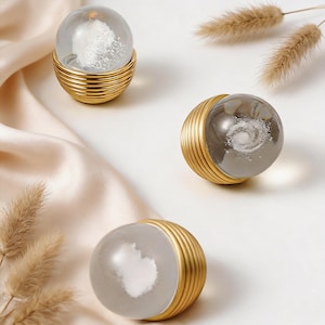 May include: Three decorative glass spheres with gold-coloured, ribbed bases. Each sphere contains a unique white design, resembling clouds or a galaxy. The spheres are arranged on a cream satin surface, with dried grass accents.