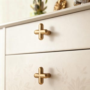May include: Close-up of a cream-colored dresser with two drawers. Each drawer features a gold-colored, cross-shaped handle. The dresser has a subtle floral pattern and a light gray top. The handles are made of metal.