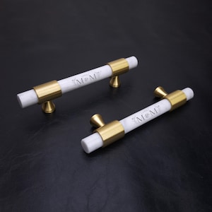 Op de afbeelding: Twee cilindrische kastgrepen met een middengedeelte van wit marmer en goudkleurige metalen eindkappen en bevestigingspunten. Elke greep is gegraveerd met de tekst M&M, omringd door een delicaat bloemmotief op het marmeren oppervlak.