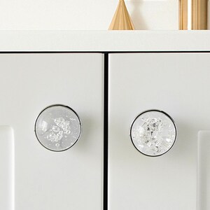 K&ouml;nnte beinhalten: Zwei transparente Schrankkn&ouml;pfe mit silberfarbenen R&auml;ndern. Die runden Kn&ouml;pfe sind mit einem strukturierten, kristallartigen Material gef&uuml;llt. Sie sind an einer wei&szlig;en Schrankt&uuml;r montiert.