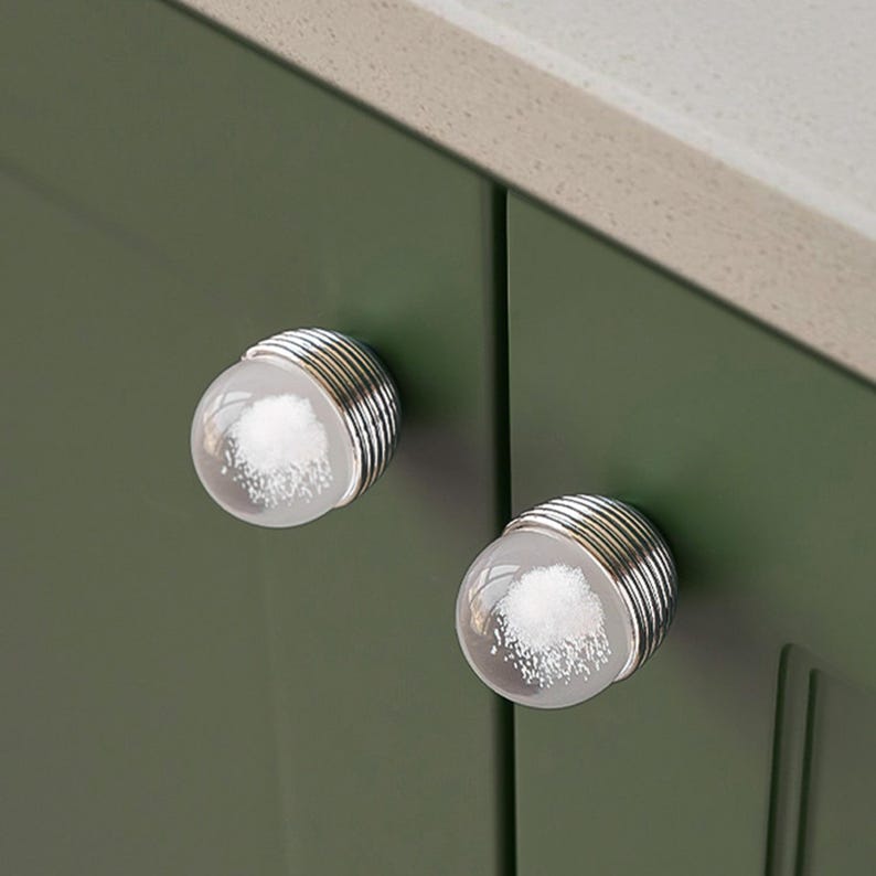 K&ouml;nnte beinhalten: Zwei transparente Glasschrankkn&ouml;pfe mit einem wei&szlig;en, wolkenartigen Design im Inneren. Die Kn&ouml;pfe haben einen silbernen, gerippten Metallsockel und sind an einer dunkelgr&uuml;nen Schrankt&uuml;r befestigt.