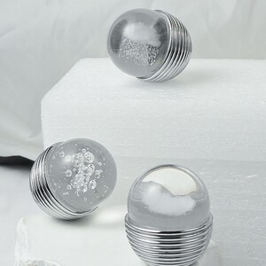 K&ouml;nnte beinhalten: Drei dekorative Kugelobjekte aus Glas und Metall. Jede Kugel hat eine klare Glaskugel mit einem wolkigen, wei&szlig;en Innenraum, die auf einem silbernen, gerippten Metallsockel sitzt. Die Kugeln sind auf einer wei&szlig;en Oberfl&auml;che angeordnet.