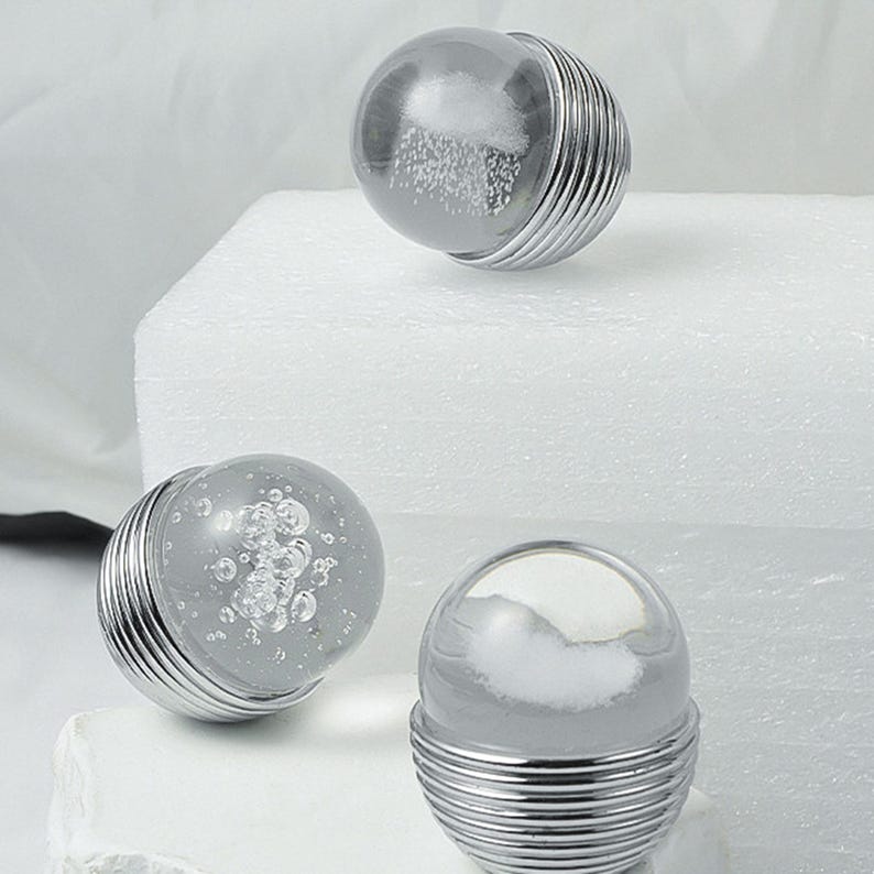 K&ouml;nnte beinhalten: Drei dekorative Kugelobjekte aus Glas und Metall. Jede Kugel hat eine klare Glaskugel mit einem wolkigen, wei&szlig;en Innenraum, die auf einem silbernen, gerippten Metallsockel sitzt. Die Kugeln sind auf einer wei&szlig;en Oberfl&auml;che angeordnet.