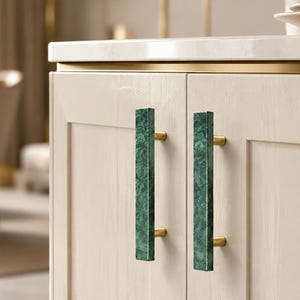 May include: Two vertical cabinet handles featuring a rectangular green marble bar with a textured, mottled surface. Each handle is attached to the light-colored wooden cabinet doors by two cylindrical gold-toned metal mounting posts.