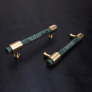 Può includere: Due maniglie cilindriche per armadietti con una parte centrale in marmo verde scuro e finiture in metallo color oro lucido sulle estremità e sui supporti. Ogni maniglia presenta l'incisione M&M in un carattere serif bianco sul marmo.