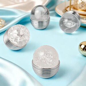 K&ouml;nnte beinhalten: Mehrere dekorative Glaskugeln mit silberfarbenen Metallsockeln sind auf einer hellblauen Oberfl&auml;che angeordnet. Die Kugeln haben verschiedene Innendesigns, darunter ein geknackter Kristalleffekt und ein blasenartiges Aussehen. Die Sockel haben ein geripptes Design.