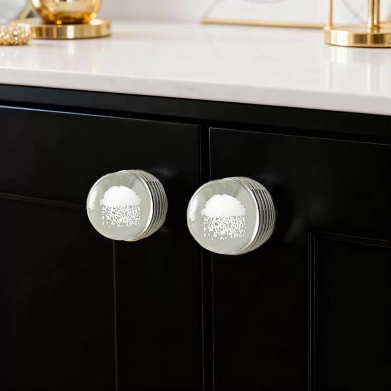 K&ouml;nnte beinhalten: Zwei transparente Glasschrankkn&ouml;pfe mit wei&szlig;em Wolkenmuster und silbernen gerippten Kanten. Die Kn&ouml;pfe sind an einer schwarzen Schrankt&uuml;r montiert. Dekoratives Zubeh&ouml;r.