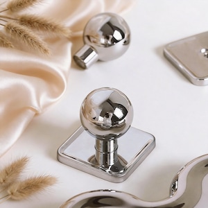 May include: Shiny, silver-colored decorative knobs with a spherical top and a square base. The knobs are arranged on a white surface with a soft, cream-colored fabric and dried ornamental grass.