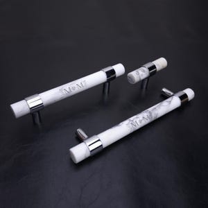 May include: Three white marble cabinet pulls with grey veining and polished chrome metal accents. Each handle features the engraved text M&M centred on the cylindrical marble bar, held in place by two chrome mounting brackets.
