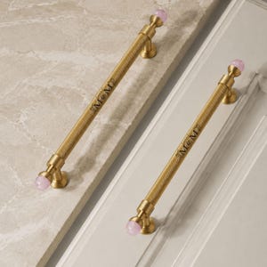 May include: Two long, cylindrical cabinet pulls made of brushed brass with a textured, knurled surface. Each handle features a small, spherical light pink stone accent at both ends. The text M&M is engraved on the centre of each metal handle.