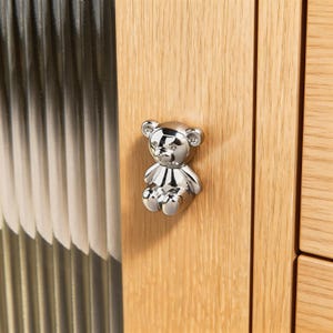 Puede incluir: Un pomo de armario plateado brillante con forma de oso de peluche está montado en una puerta de armario de madera clara. El pomo es un diseño tridimensional detallado de un oso sentado. La puerta tiene vetas de madera verticales y un panel de vidrio a la izquierda.