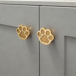 May include: Two gold-colored paw print cabinet knobs mounted on a gray cabinet door. The knobs are shaped like a dog's paw, with five circular pads and a small screw for mounting. The cabinet is painted a solid gray color.