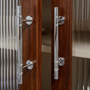 May include: Two polished chrome cabinet door handles with a bamboo-like design are mounted on a dark wood cabinet. The handles are long and cylindrical, with decorative rings and circular mounting plates.