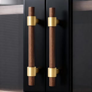 May include: Two cabinet door handles. Each handle features a dark brown, cylindrical wooden grip with a gold-coloured metal band in the centre and at the base. The handles are mounted on a black cabinet door.