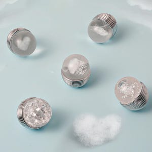 K&ouml;nnte beinhalten: F&uuml;nf klare, kugelf&ouml;rmige Objekte mit silbernen Metallsockeln und -oberteilen. Jede Kugel enth&auml;lt ein anderes wei&szlig;es Design, darunter ein Herz und wolkenartige Formen. Die Objekte sind auf einer hellblauen Oberfl&auml;che angeordnet.