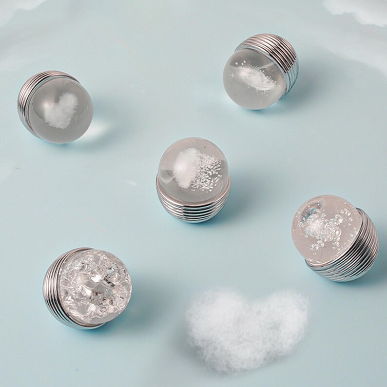 K&ouml;nnte beinhalten: F&uuml;nf klare, kugelf&ouml;rmige Objekte mit silbernen Metallsockeln und -oberteilen. Jede Kugel enth&auml;lt ein anderes wei&szlig;es Design, darunter ein Herz und wolkenartige Formen. Die Objekte sind auf einer hellblauen Oberfl&auml;che angeordnet.