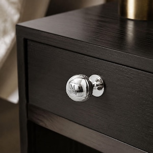 May include: A polished silver spherical drawer knob attached to a dark wood drawer. The knob features a circular engraving with the letters M and M separated by an ampersand, framed by delicate leaf motifs on the top and bottom of the lettering.
