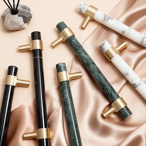 Op de afbeelding: Een verzameling kastgrepen in verschillende stijlen. Sommige zijn zwart, andere wit met grijze adering en sommige groen met donkerdere groene adering. Allemaal hebben ze goudkleurige metalen accenten. De grepen zijn gerangschikt op een satijnachtig oppervlak.