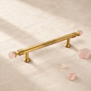 May include: A brushed gold cabinet handle featuring a cylindrical bar with ribbed ends and spherical pink quartz finials. The centre of the bar is engraved with the text M&M surrounded by delicate floral line art, accompanied by loose pink quartz stones.