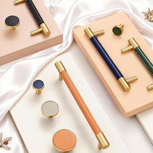 May include: A collection of cabinet hardware, including gold-toned handles and knobs. The handles feature leather-wrapped grips in black, blue, green, and orange. The knobs are round with matching leather and gold accents. The hardware is displayed on a white and peach background.