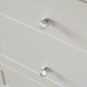 K&ouml;nnte beinhalten: Nahaufnahme eines wei&szlig;en Schranks mit zwei durchsichtigen, kugelf&ouml;rmigen Kn&ouml;pfen. Jeder Knopf hat einen silbernen Metallsockel mit horizontalen Linien. Der Schrank hat horizontale, gerillte Paneele. Die Kn&ouml;pfe sind gleichm&auml;&szlig;ig angeordnet.