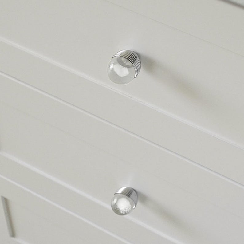 K&ouml;nnte beinhalten: Nahaufnahme eines wei&szlig;en Schranks mit zwei durchsichtigen, kugelf&ouml;rmigen Kn&ouml;pfen. Jeder Knopf hat einen silbernen Metallsockel mit horizontalen Linien. Der Schrank hat horizontale, gerillte Paneele. Die Kn&ouml;pfe sind gleichm&auml;&szlig;ig angeordnet.