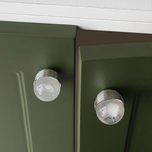 K&ouml;nnte beinhalten: Zwei Schrankkn&ouml;pfe mit klarem, kugelf&ouml;rmigem Design. Jeder Knopf hat einen silbernen, geriffelten Sockel und ist an einer dunkelgr&uuml;nen Schrankt&uuml;r befestigt. Die Kn&ouml;pfe verleihen dem Schrank eine dekorative Note.