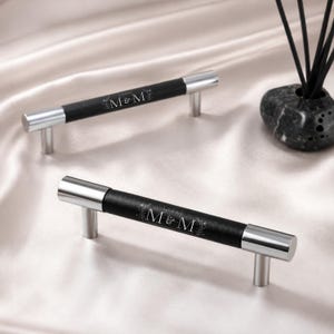 May include: Two cabinet pulls featuring a black cylindrical center with a silver metallic finish on the ends and mounting posts. The black center is engraved with the text M&M surrounded by a delicate floral wreath design in white.